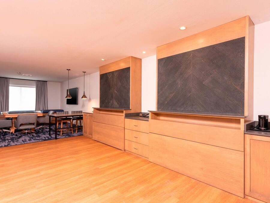 A modern hotel room with a wooden floor, a dining area with a table and chairs, a large flat-screen TV, and built-in cabinets.