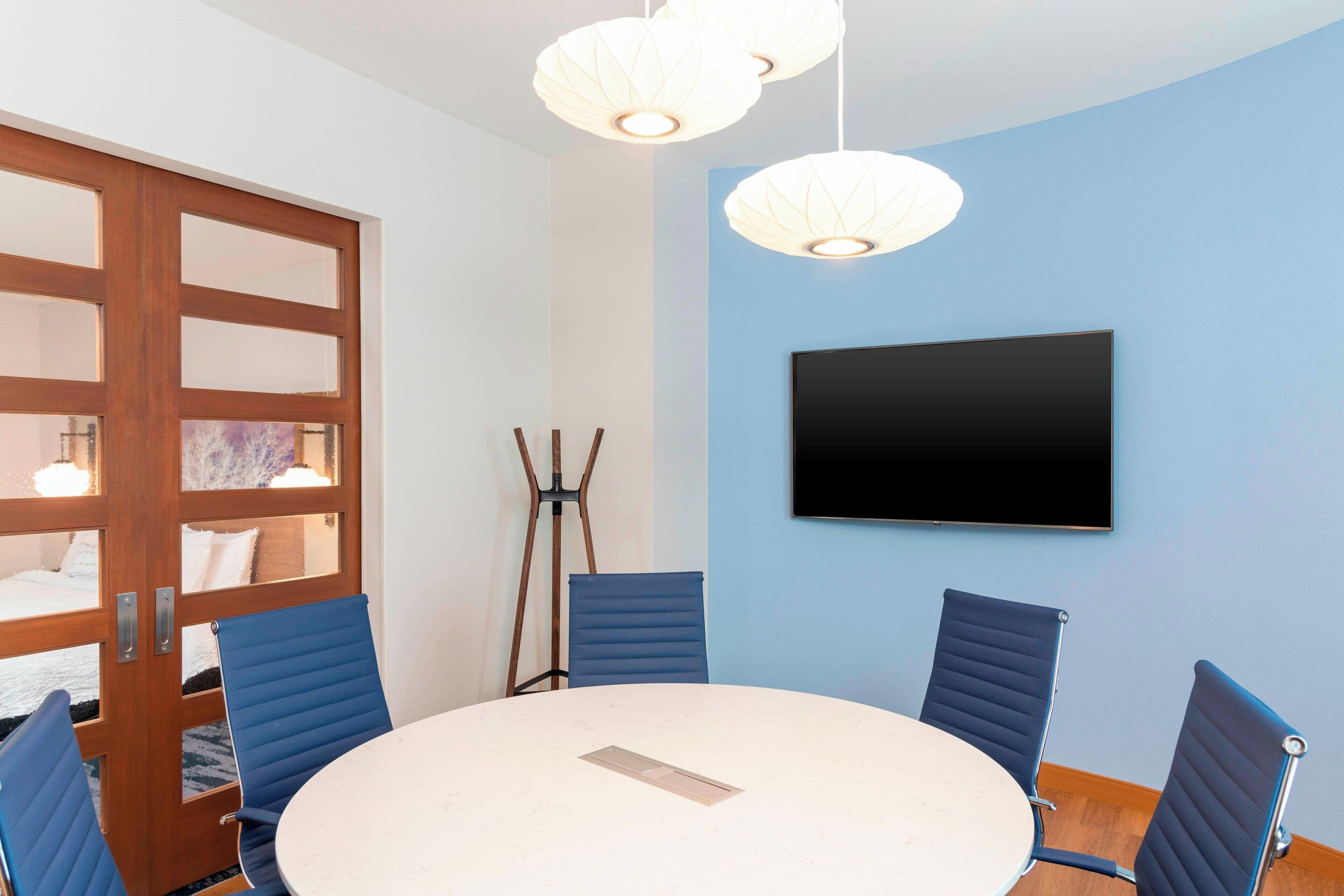 A conference room with a round table, four blue chairs, a flat-screen TV on the wall, and a wooden door with glass panels.