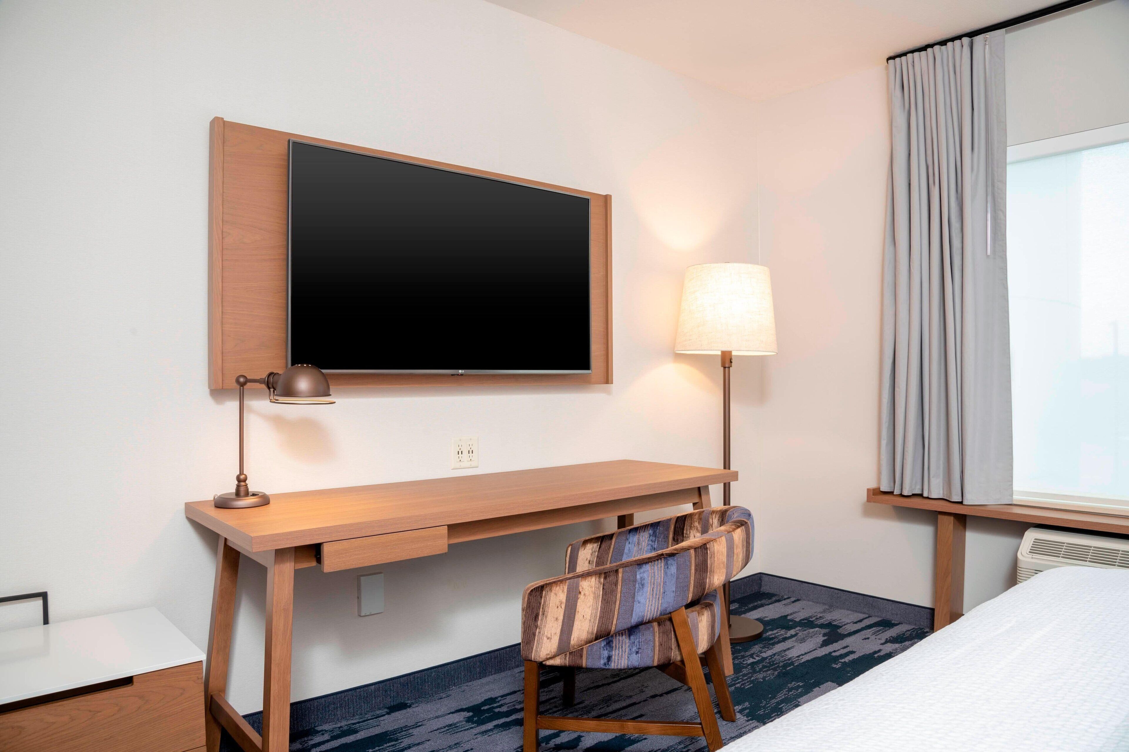 A hotel room with a wooden desk, a television, a lamp, a chair, and a bed.