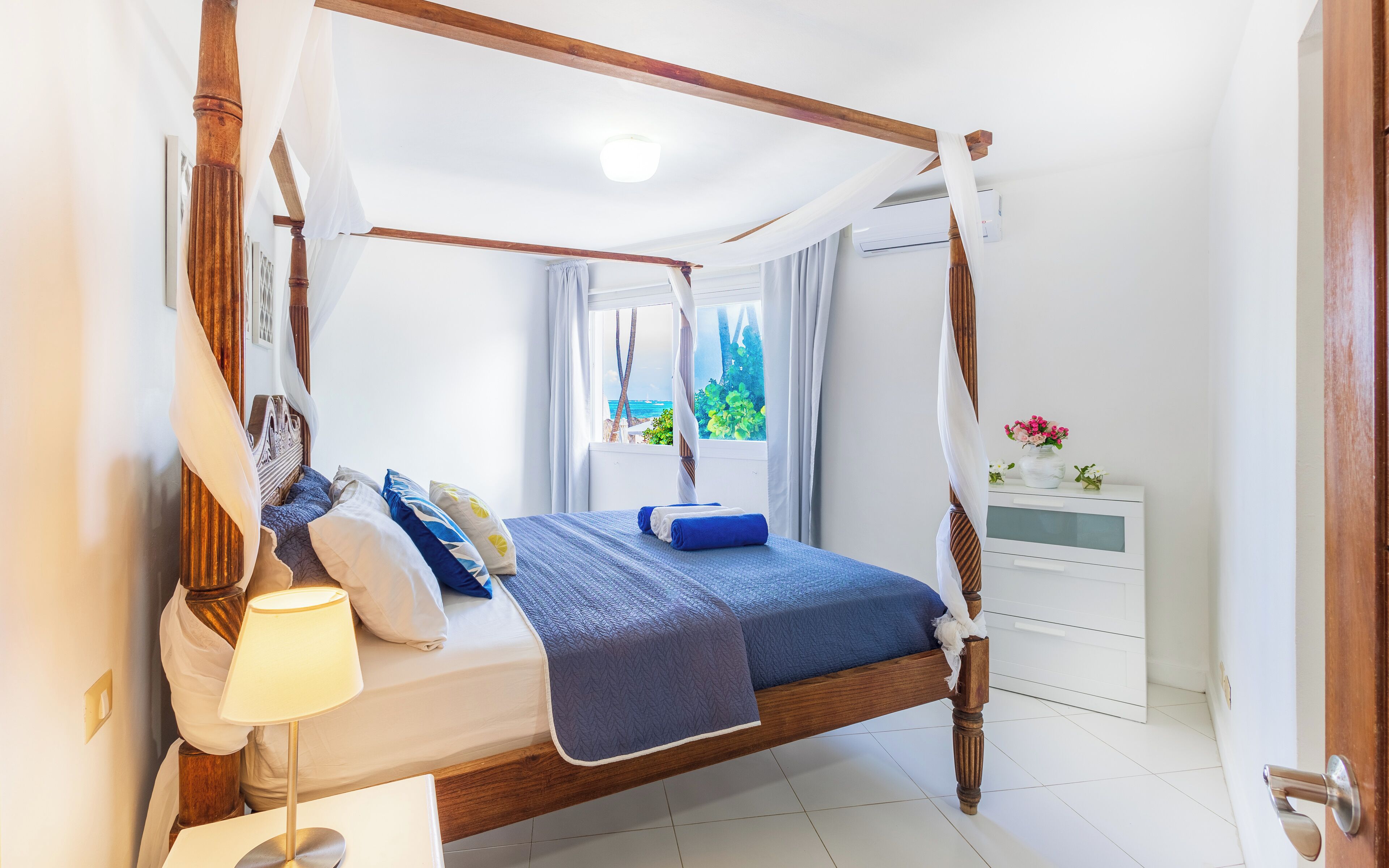 Una cama con dosel, una mesita de noche con lámpara, y vista al mar por la ventana.