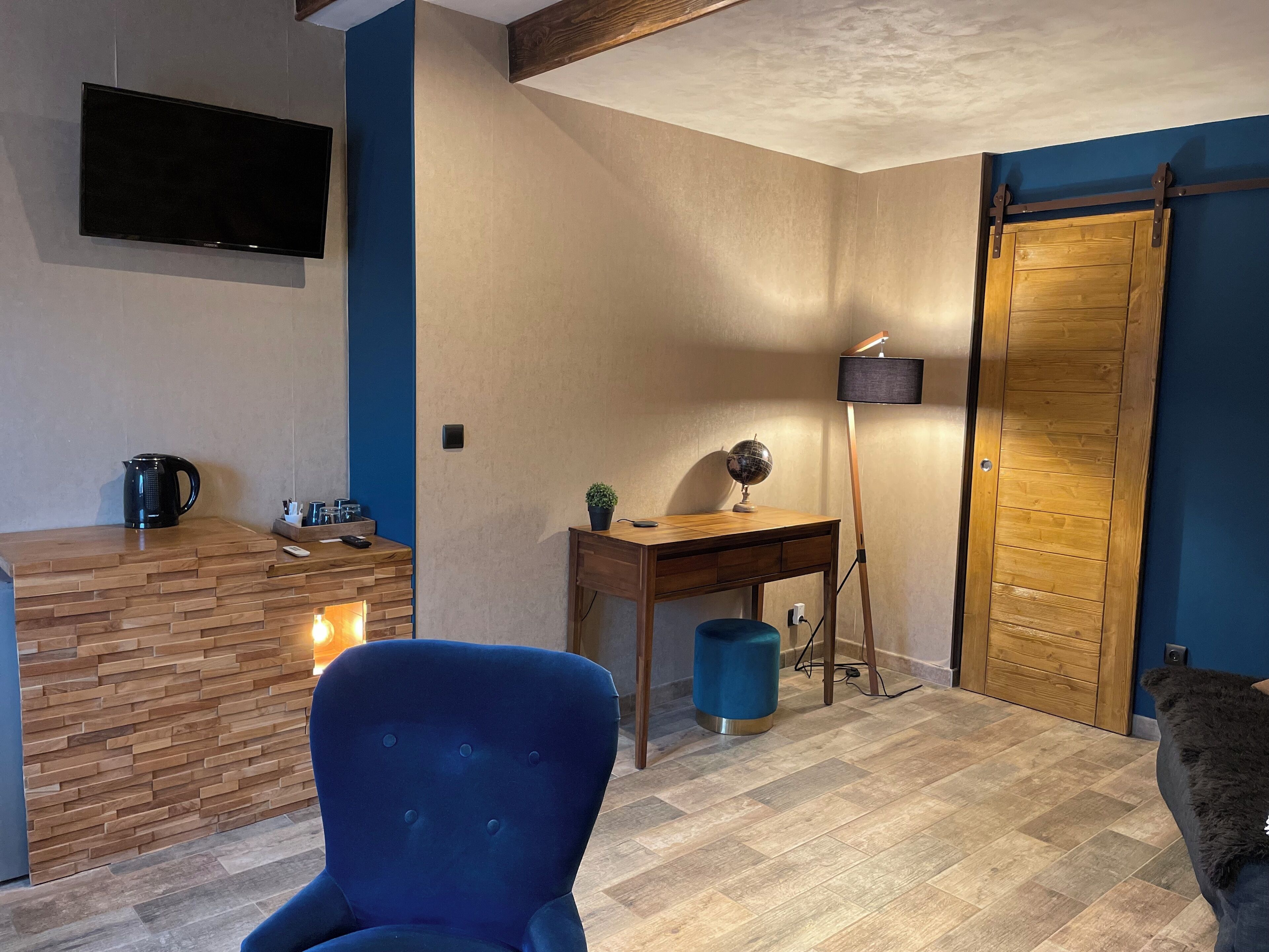 Ein modernes Wohnzimmer mit Couchtisch aus Holz, einem blauen Sessel, einem Schreibtisch mit Globus und Lampe sowie einem an der Wand befestigten Fernseher.