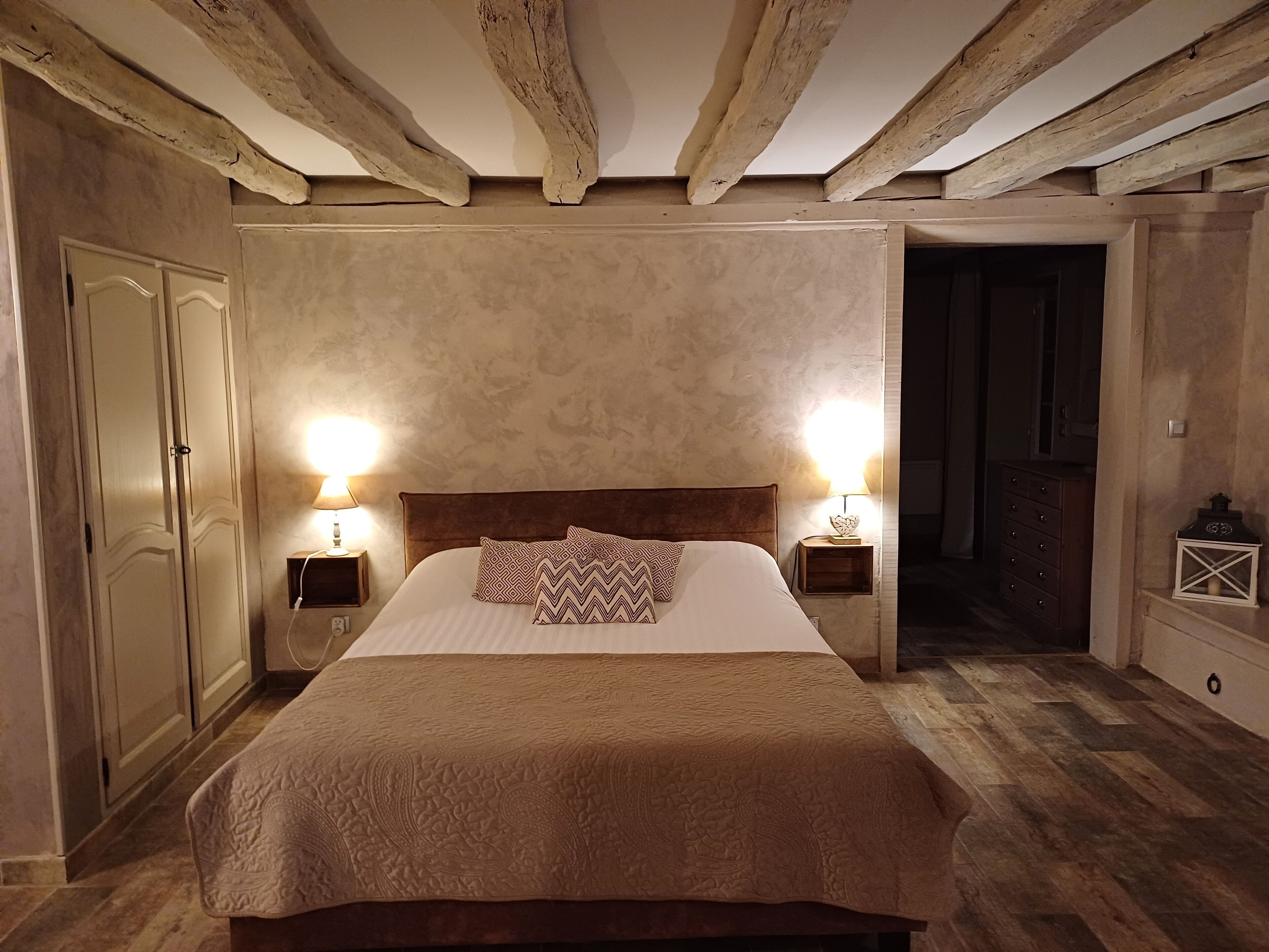 A bedroom with a large bed, wooden beams on the ceiling, two bedside lamps, a wooden dresser, and a wardrobe.