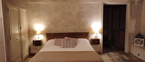 A bedroom with a large bed, wooden beams on the ceiling, two bedside lamps, a wooden dresser, and a wardrobe.