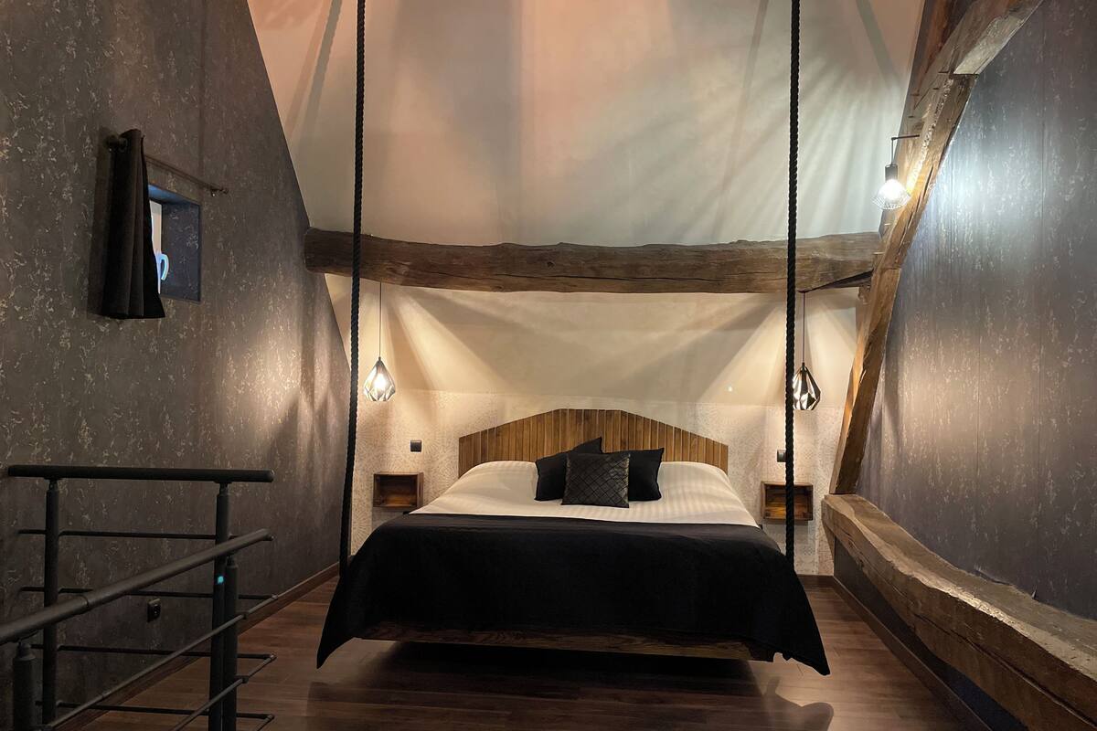 Ein Schlafzimmer mit hölzernem Kopfteil, einem Bett mit schwarzer Bettwäsche, einer Pendelleuchte und einem Holzboden.