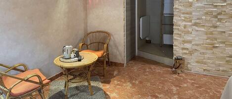 A cozy room with a wicker chair, a small round table with a kettle and cups, and a stone wall.