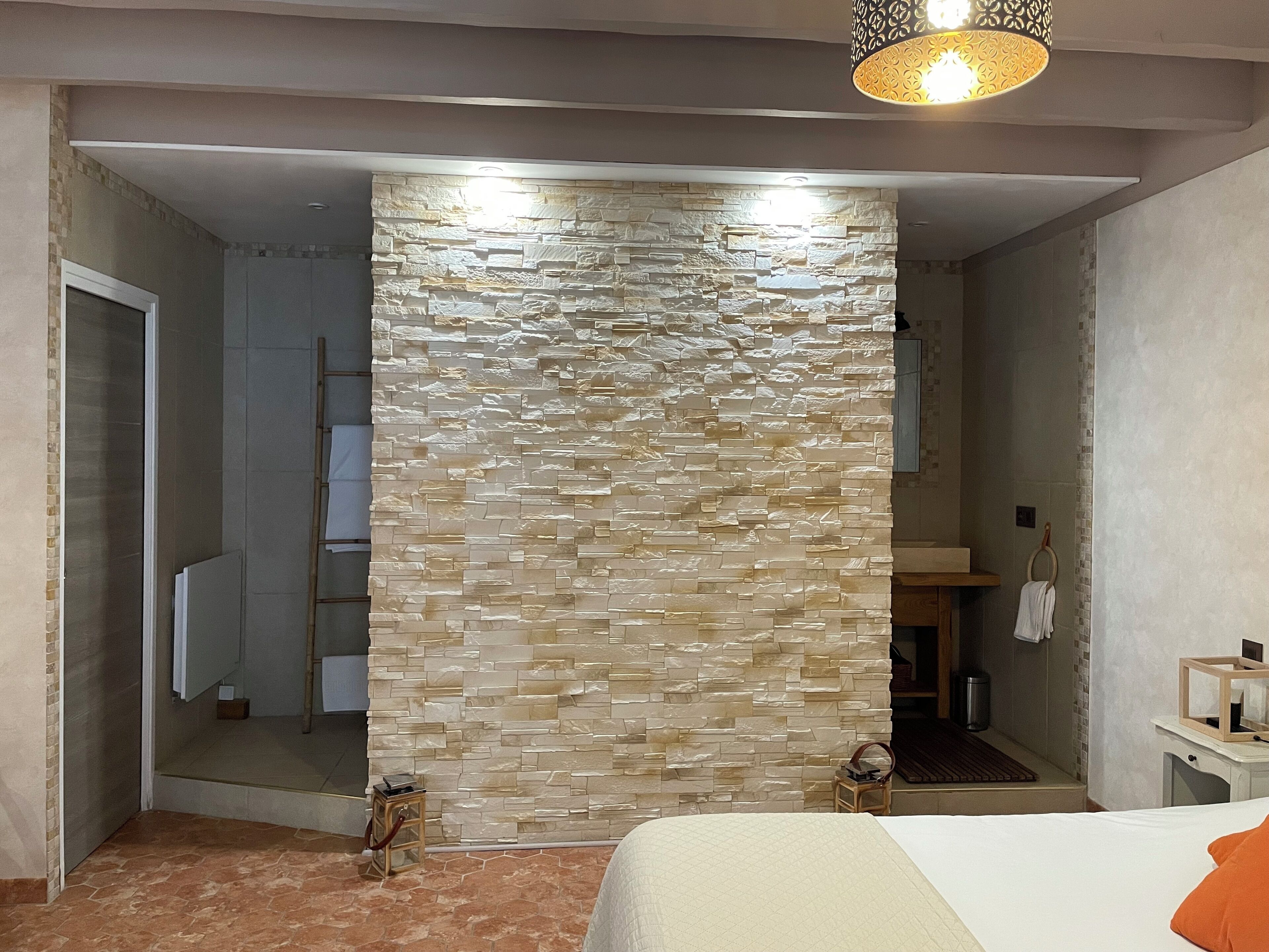 A hotel room with a stone wall, a bed with an orange pillow, a wooden nightstand, and a hanging light fixture.