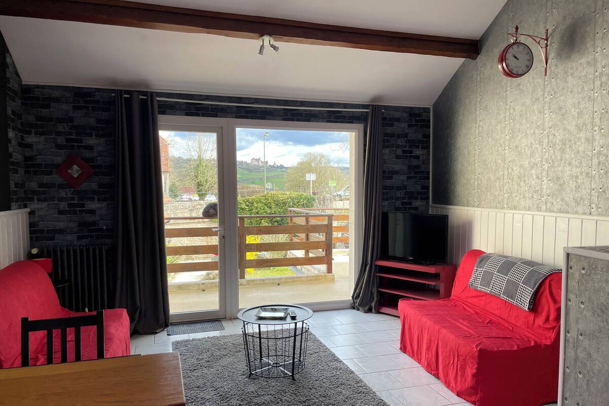 Ein Wohnzimmer mit einem roten Sofa, einem Holztisch, einem Fernseher und Blick ins Freie durch eine Schiebetür.