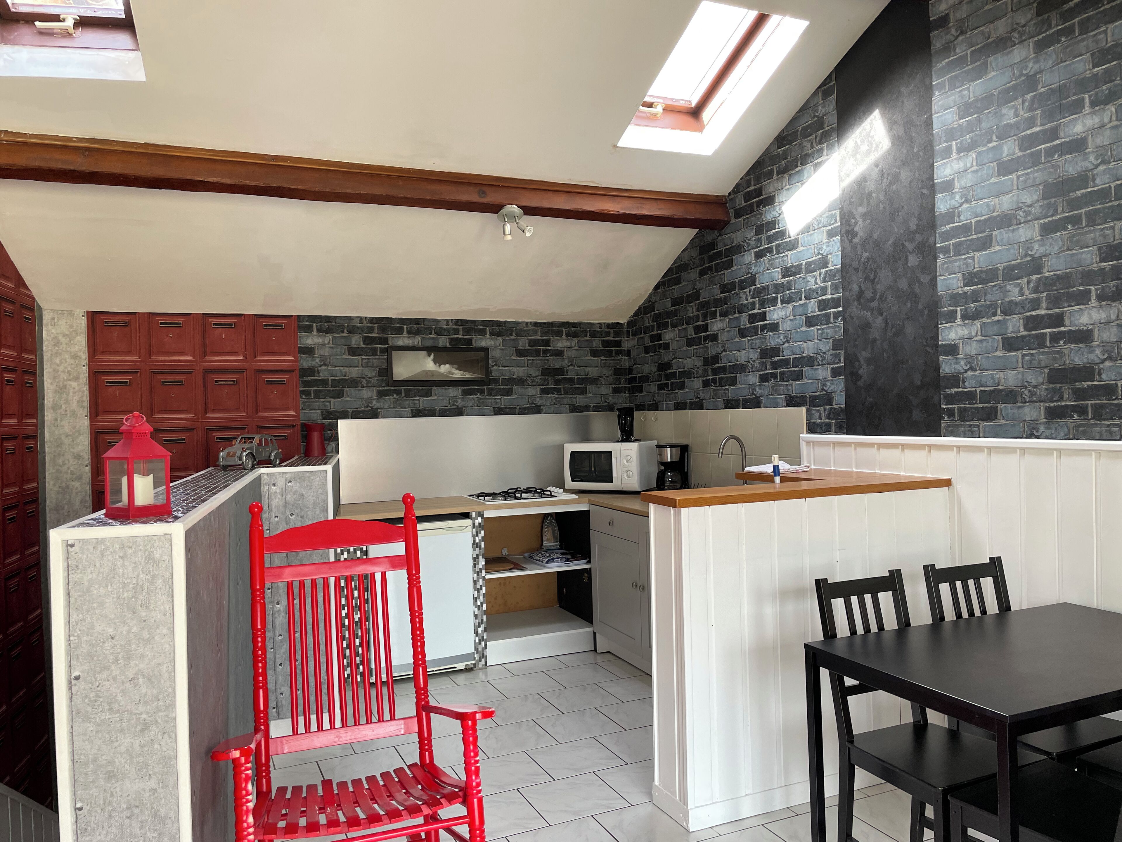 A compact kitchen with a gas stove, microwave, and sink. There is a red chair and a dining table with chairs. The kitchen has a sloped ceiling with skylights and a brick wall.