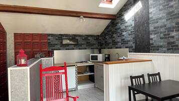 A compact kitchen with a gas stove, microwave, and sink. There is a red chair and a dining table with chairs. The kitchen has a sloped ceiling with skylights and a brick wall.