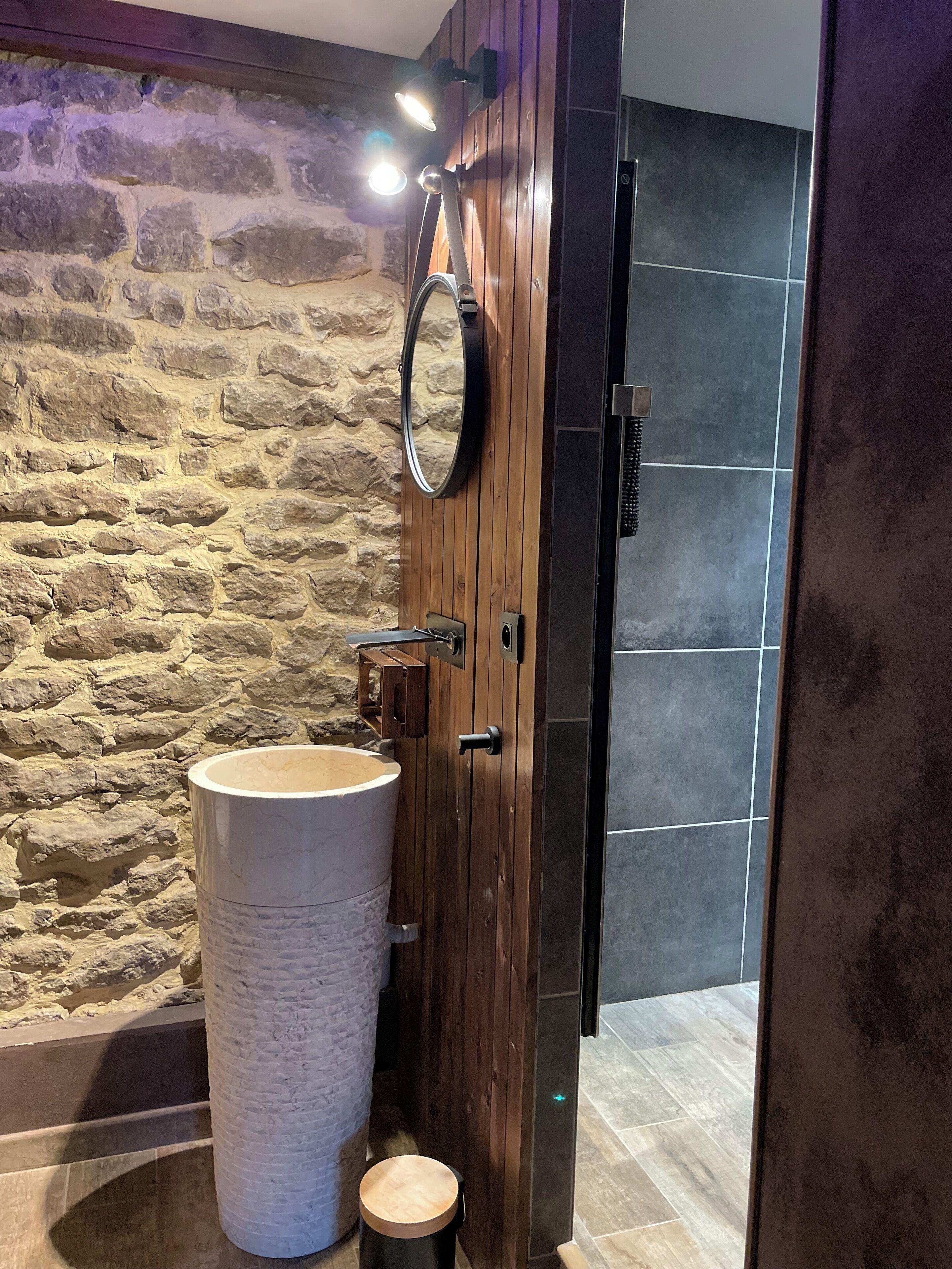 Une salle de bain avec un mur en pierre, une porte en bois, un grand miroir rond et un grand lavabo.