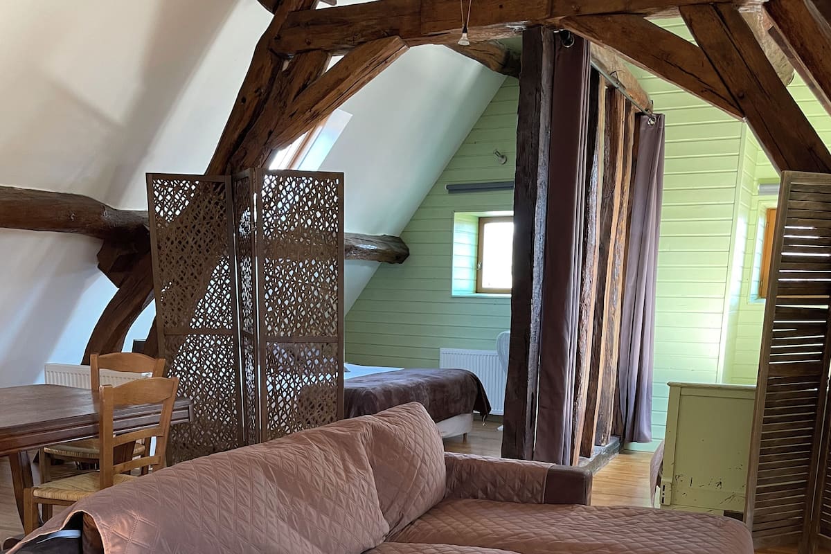Ein Loft-Zimmer mit freiliegenden Holzbalken, einem großen braunen Ecksofa, einem Esstisch mit Stühlen und einem gemusterten Teppich.