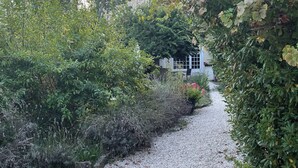Un sentiero sterrato conduce a una casa in pietra circondata da vegetazione lussureggiante e piante fiorite.