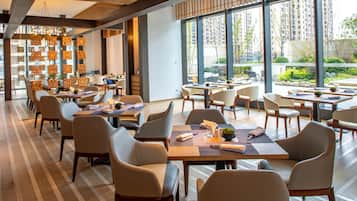 A modern dining area with multiple tables and chairs, large windows offering views of a cityscape, and decorative wall panels.