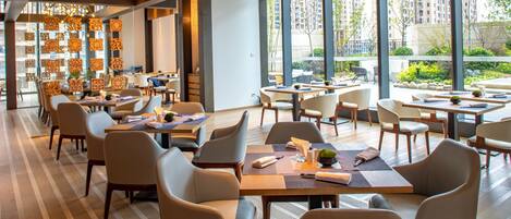 A modern dining area with multiple tables and chairs, large windows offering views of a cityscape, and decorative wall panels.