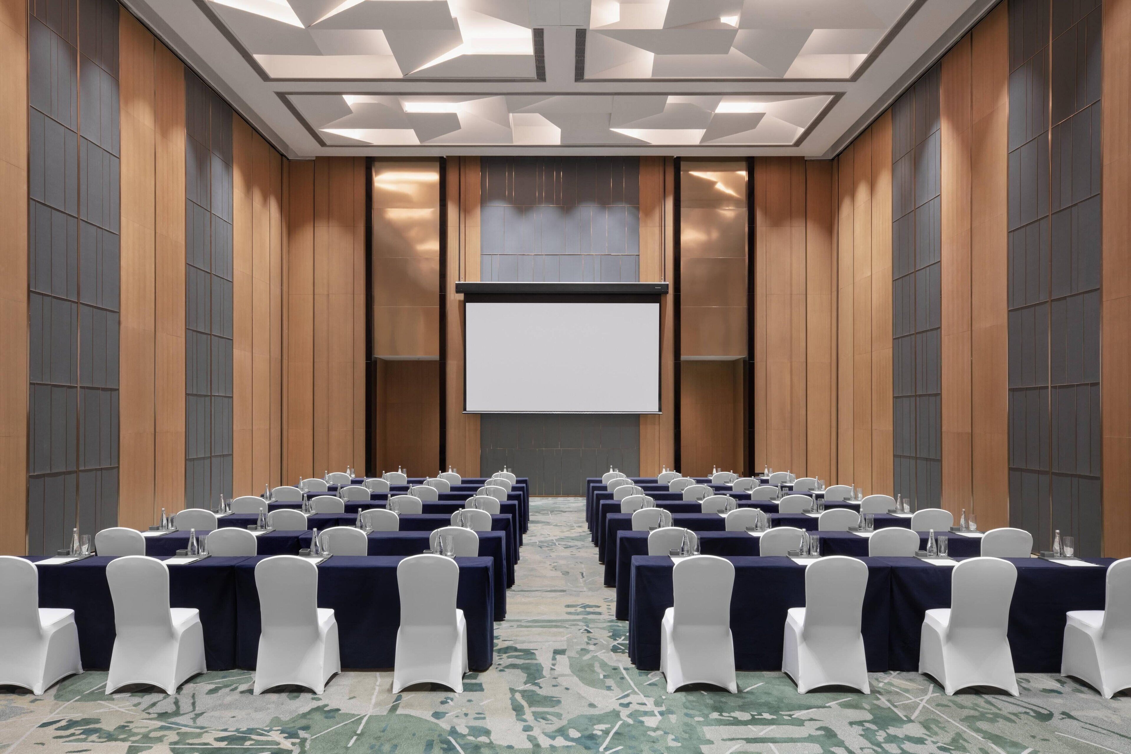 A conference room with rows of tables, white chairs, and a projector screen.