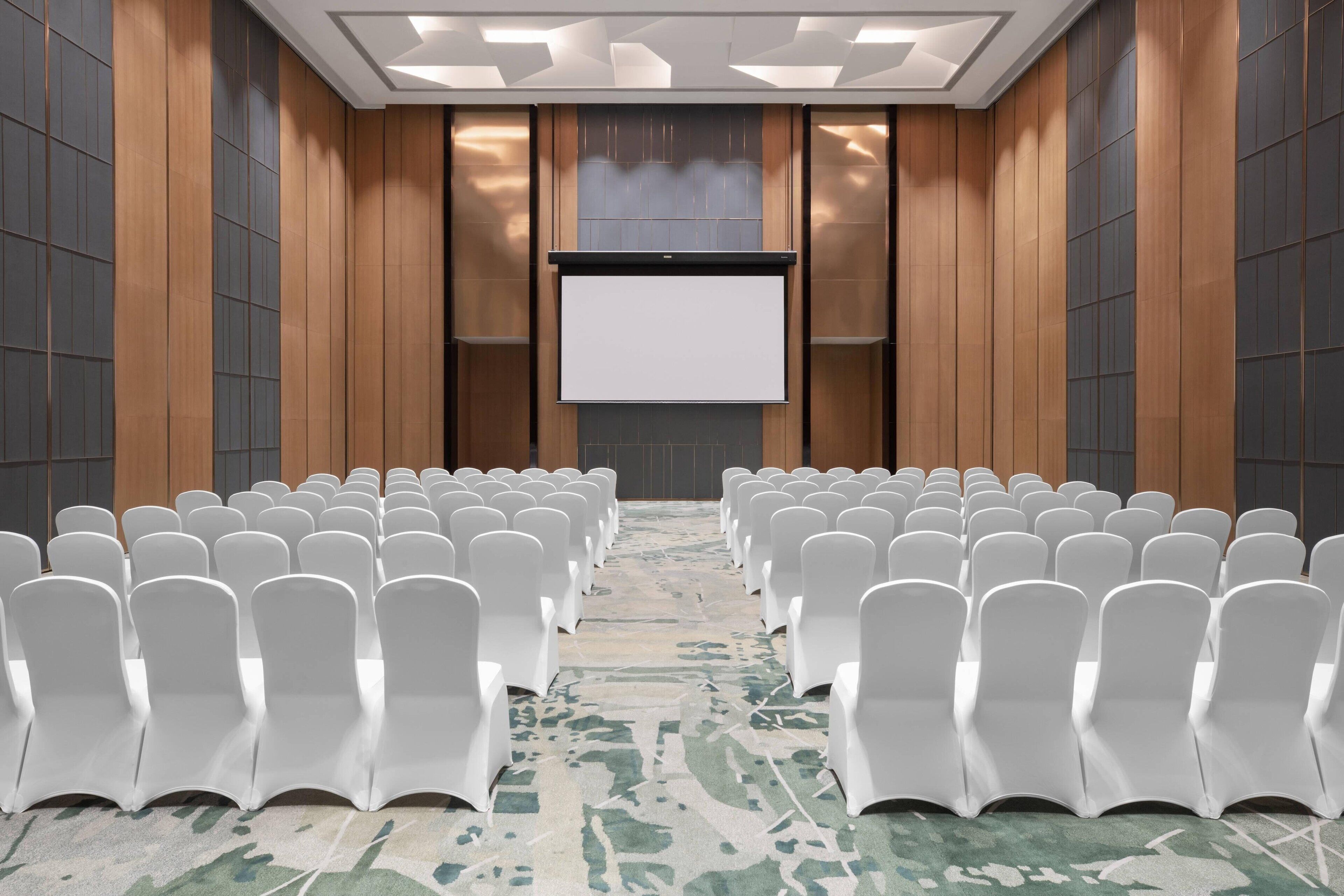 Een congreszaal met rijen witte stoelen, een groot scherm aan de voorkant en een kamerbreed tapijt met een patroon.