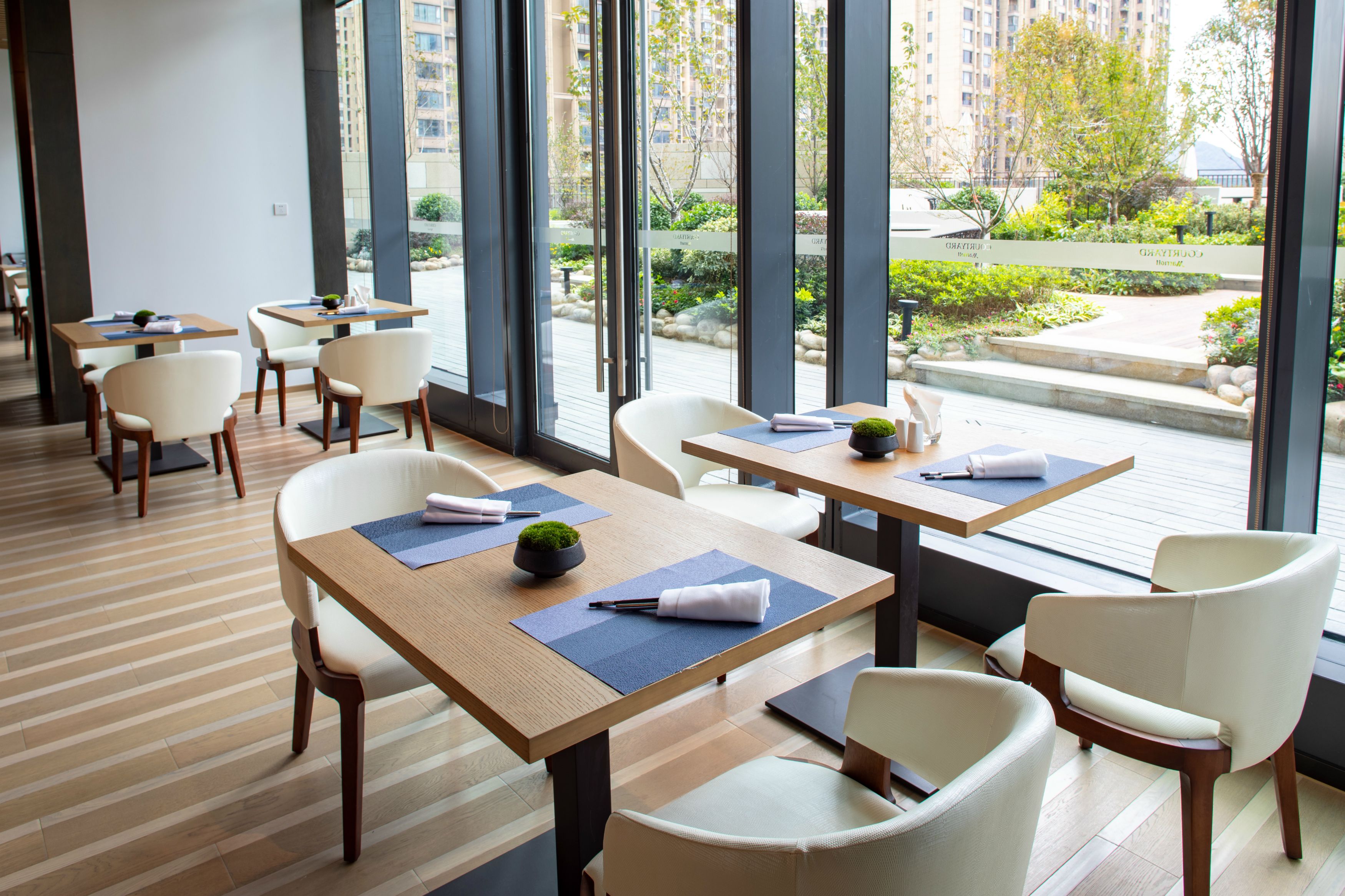 A modern hotel lobby with wooden flooring, white chairs, and tables with blue placemats. Large windows offer views of a landscaped garden and buildings.