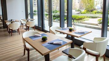A modern hotel lobby with wooden flooring, white chairs, and tables with blue placemats. Large windows offer views of a landscaped garden and buildings.