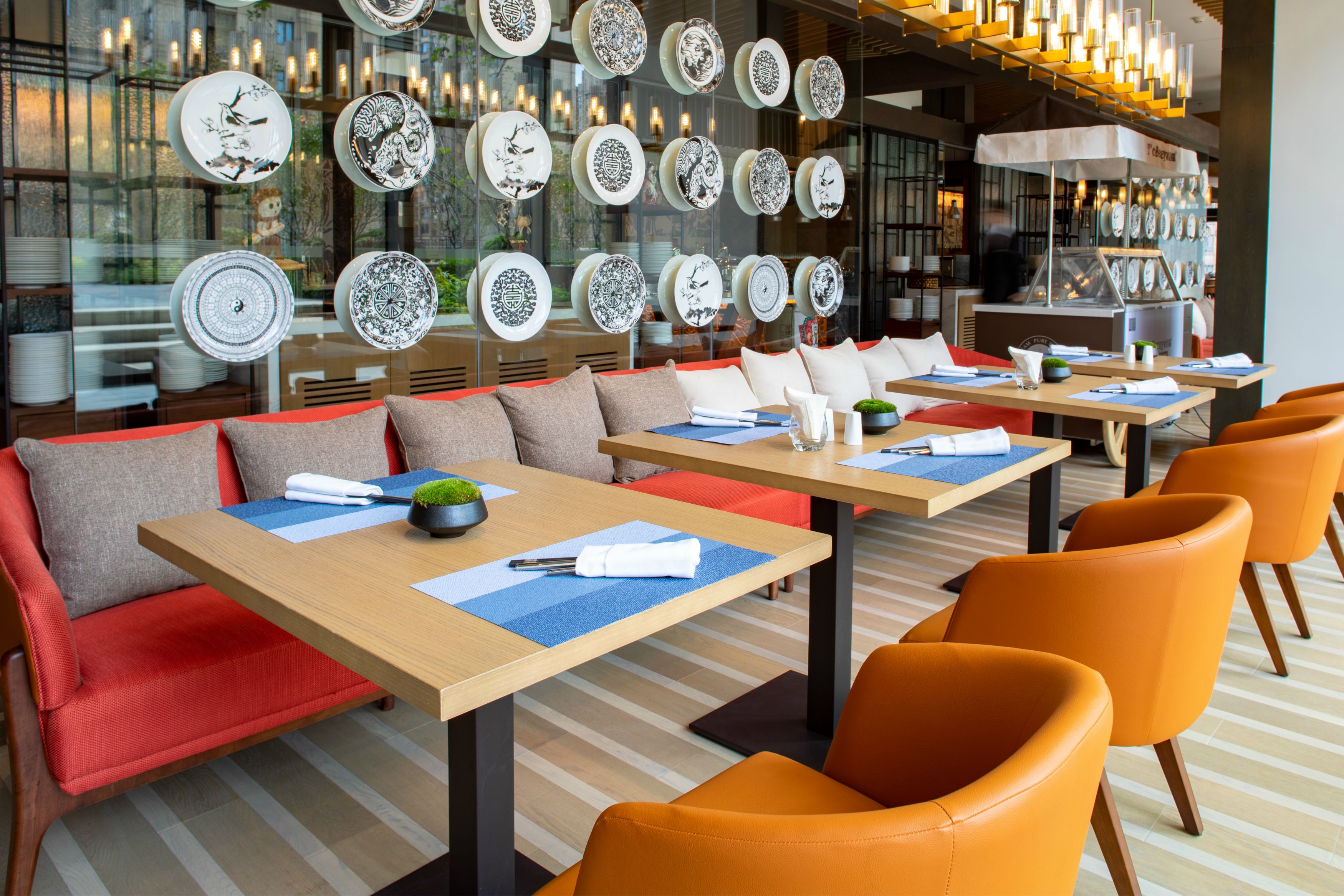 Un restaurante moderno con varias mesas y sillas anaranjadas, que presenta una pared de platos decorativos y una partición de vidrio.