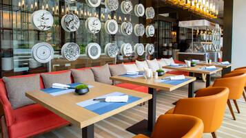 Een modern restaurant met meerdere tafels en oranje stoelen, met een wand decoratieve borden en een glazen scheiding.