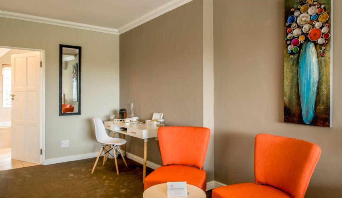 A hotel room with a desk, two orange chairs, a white chair, a painting on the wall, and a door leading to another room.
