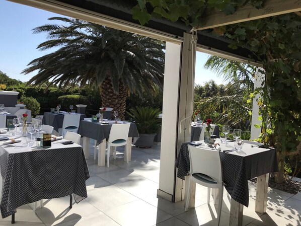 Een buitenterras met tafels voor twee personen, beschaduwd door parasols en omgeven door groen.