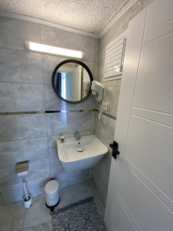 A bathroom with a white sink, a round mirror, a hairdryer, a toilet, and a floor mat.