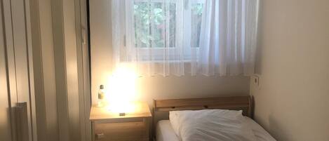 Une petite chambre à coucher, bien rangée, avec un lit simple, une table de chevet avec une lampe et une fenêtre ornée de rideaux légers.