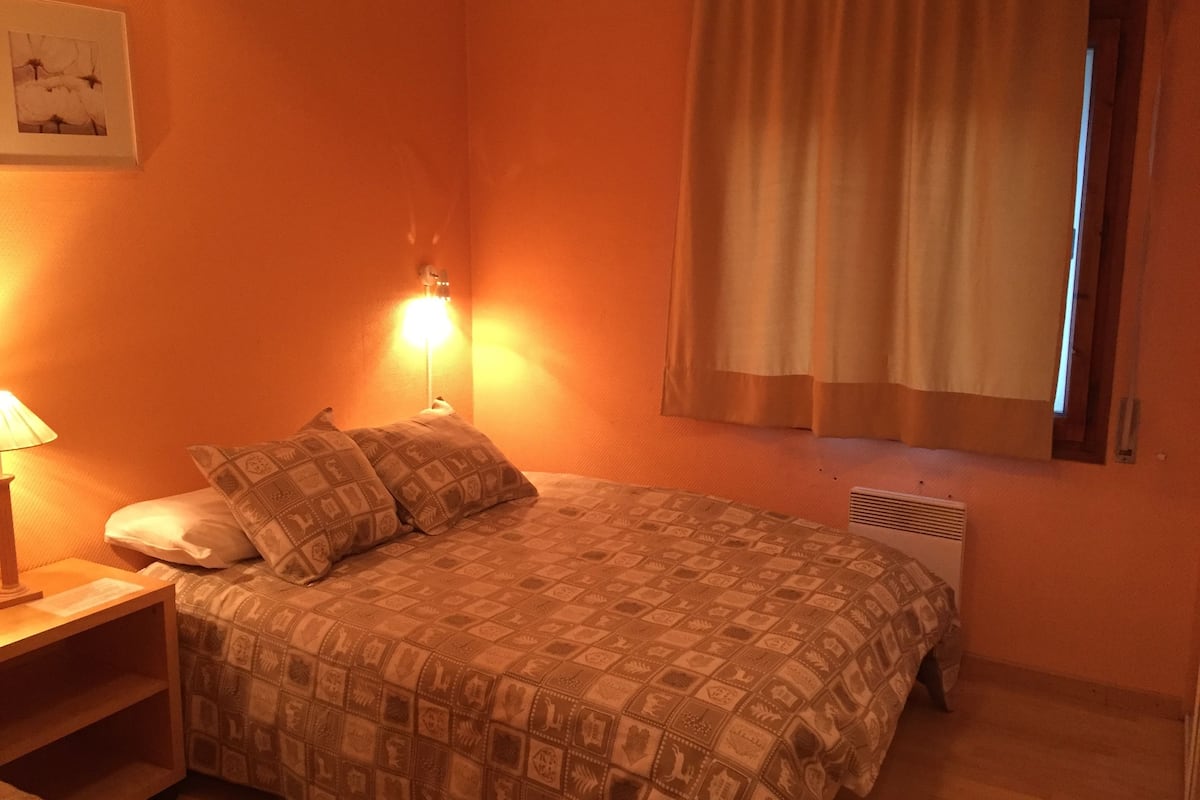 A bedroom with a bed, a nightstand, a lamp, a wall-mounted light, a window with curtains, and a radiator.