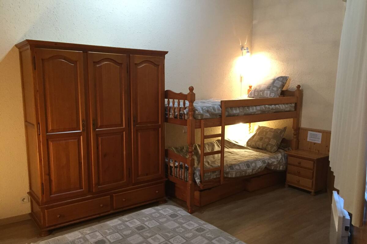 A room with a bunk bed, a wooden wardrobe, a bedside table, and a patterned rug.