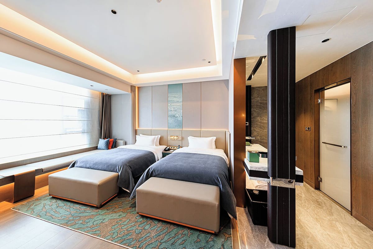 Una habitación de hotel moderna con dos camas, un ventanal grande, un espacio de descanso y un baño con ducha e inodoro.