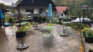 An outdoor patio area with tables and chairs, a building with a sloped roof, and a forested hillside in the background. - Sporthostel Rössle (Schluchsee)