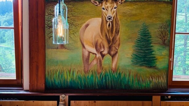 A dining area with a table set for two, a painting of a deer on the wall, and a view of the outdoors through the windows. - Sporthostel Rössle (Schluchsee)