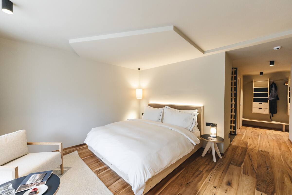 A modern bedroom with a wooden floor, a bed with white bedding, a bedside table with a lamp, and a chair.
