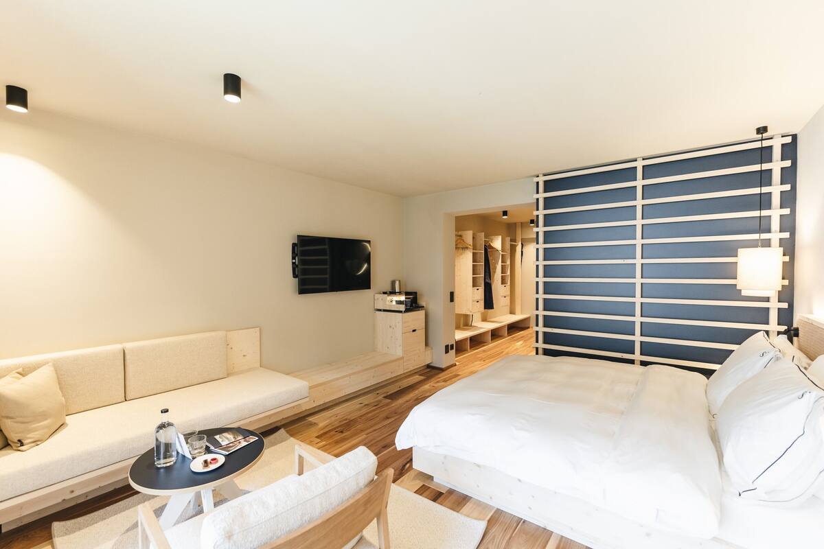 A modern hotel room with a bed, a sofa, a small table with a bottle and glasses, and a TV on the wall.