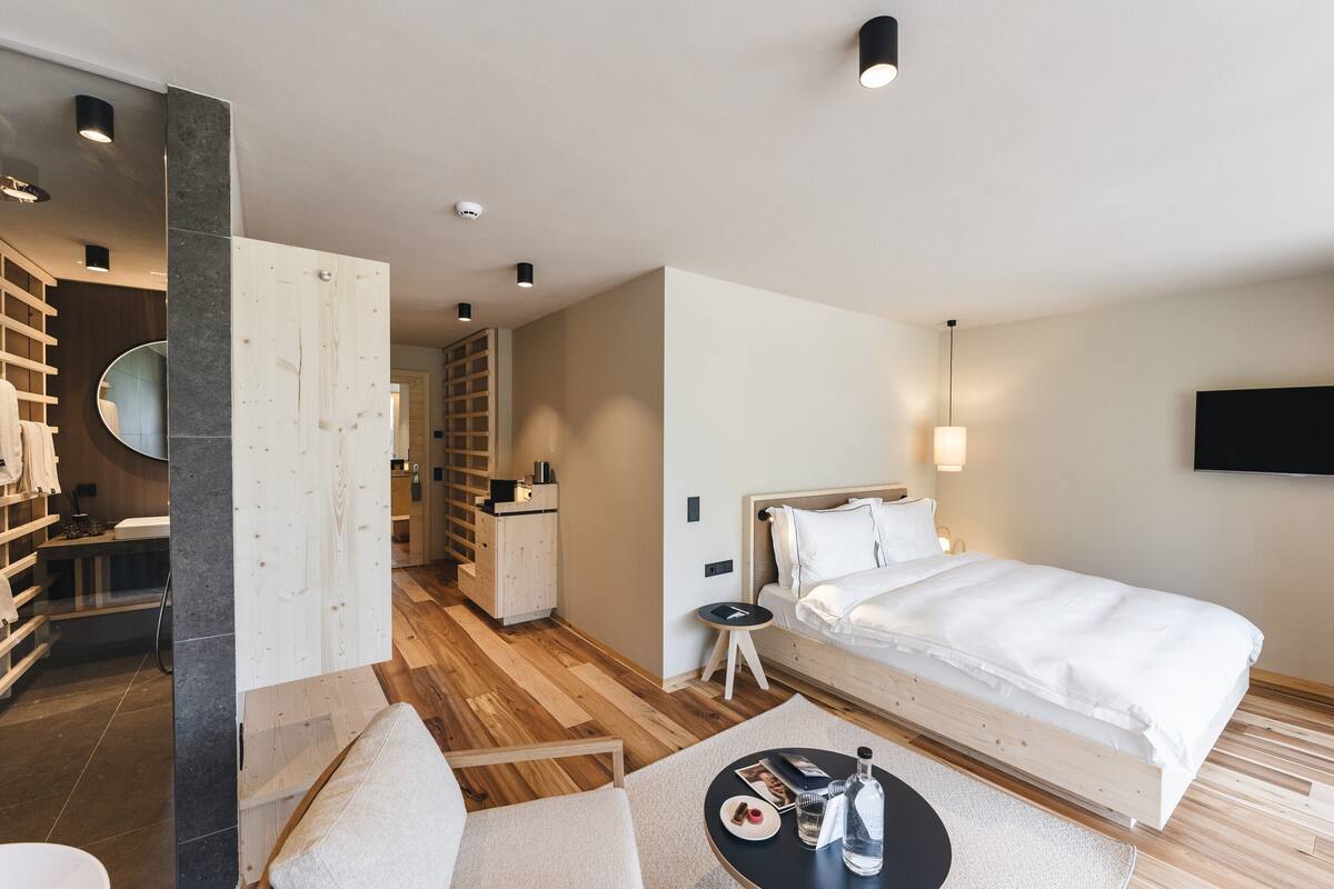 A modern hotel room with a wooden floor, a bed with white bedding, a small round table with a bottle and a plate, and a TV mounted on the wall.