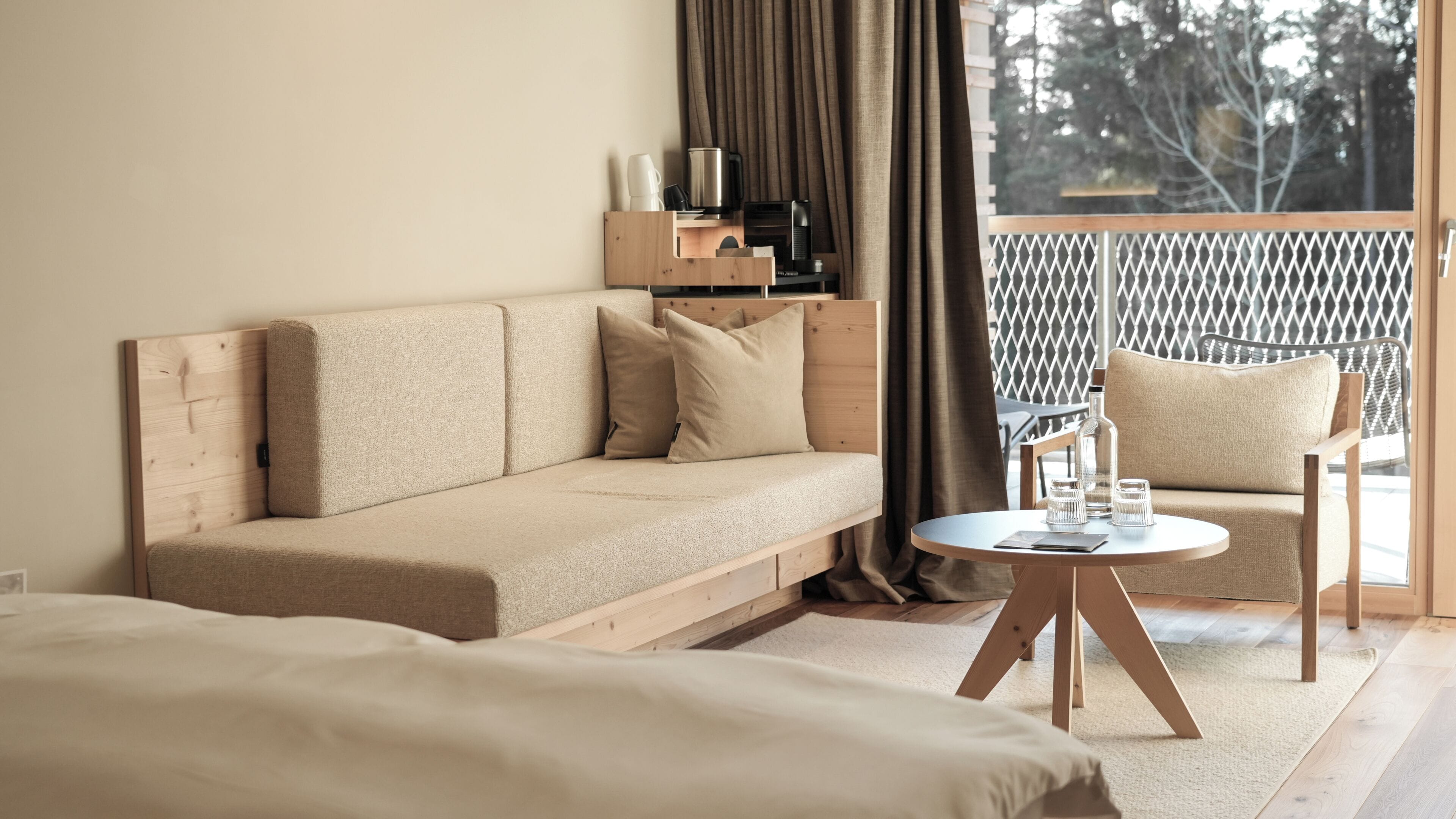 A modern hotel room with a sofa, a small round table with glasses, a chair, and a balcony with a lattice railing.