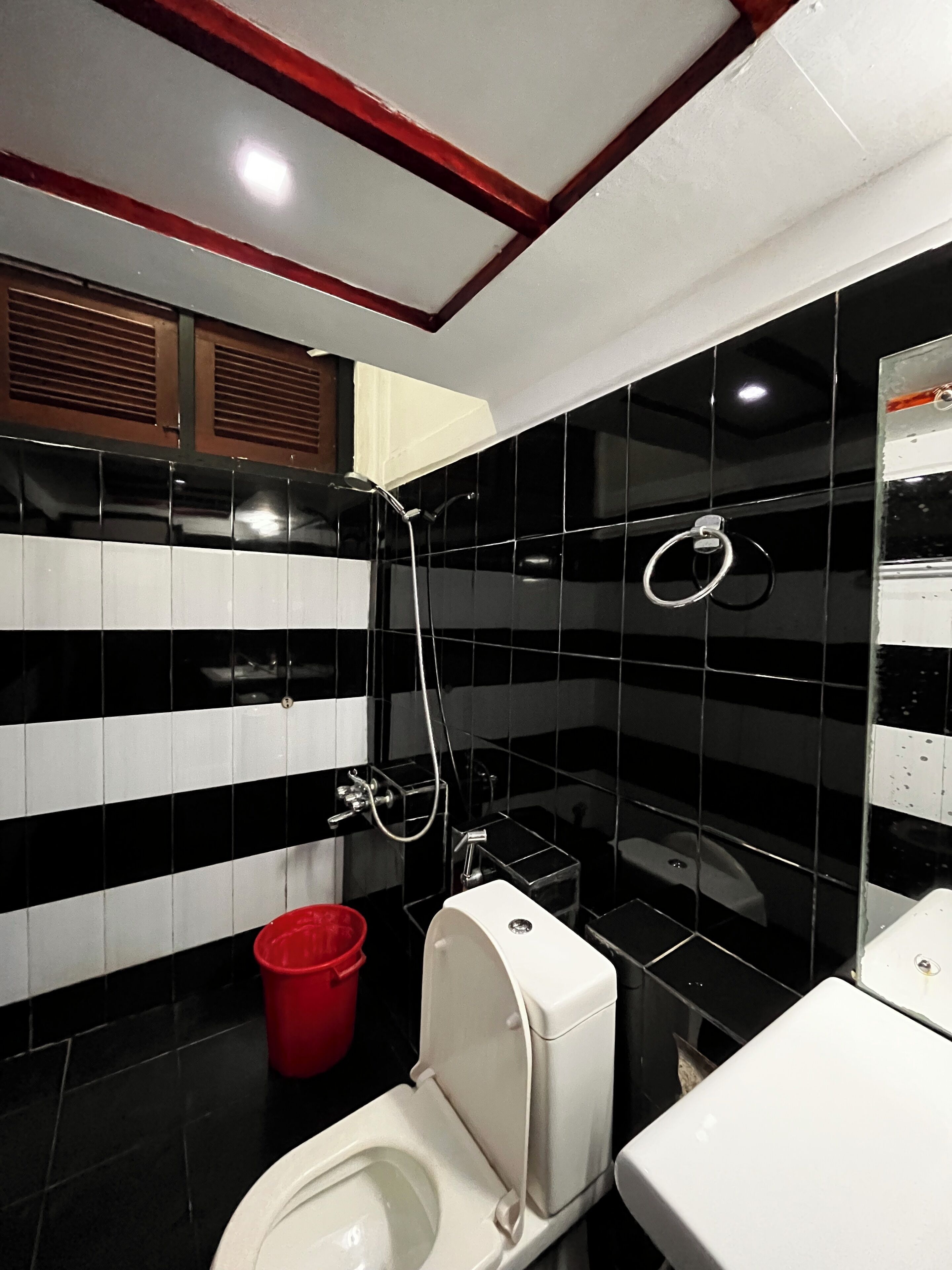 Un baño con paredes de azulejos blanco y negro, un inodoro blanco, un cubo rojo y una zona de ducha.