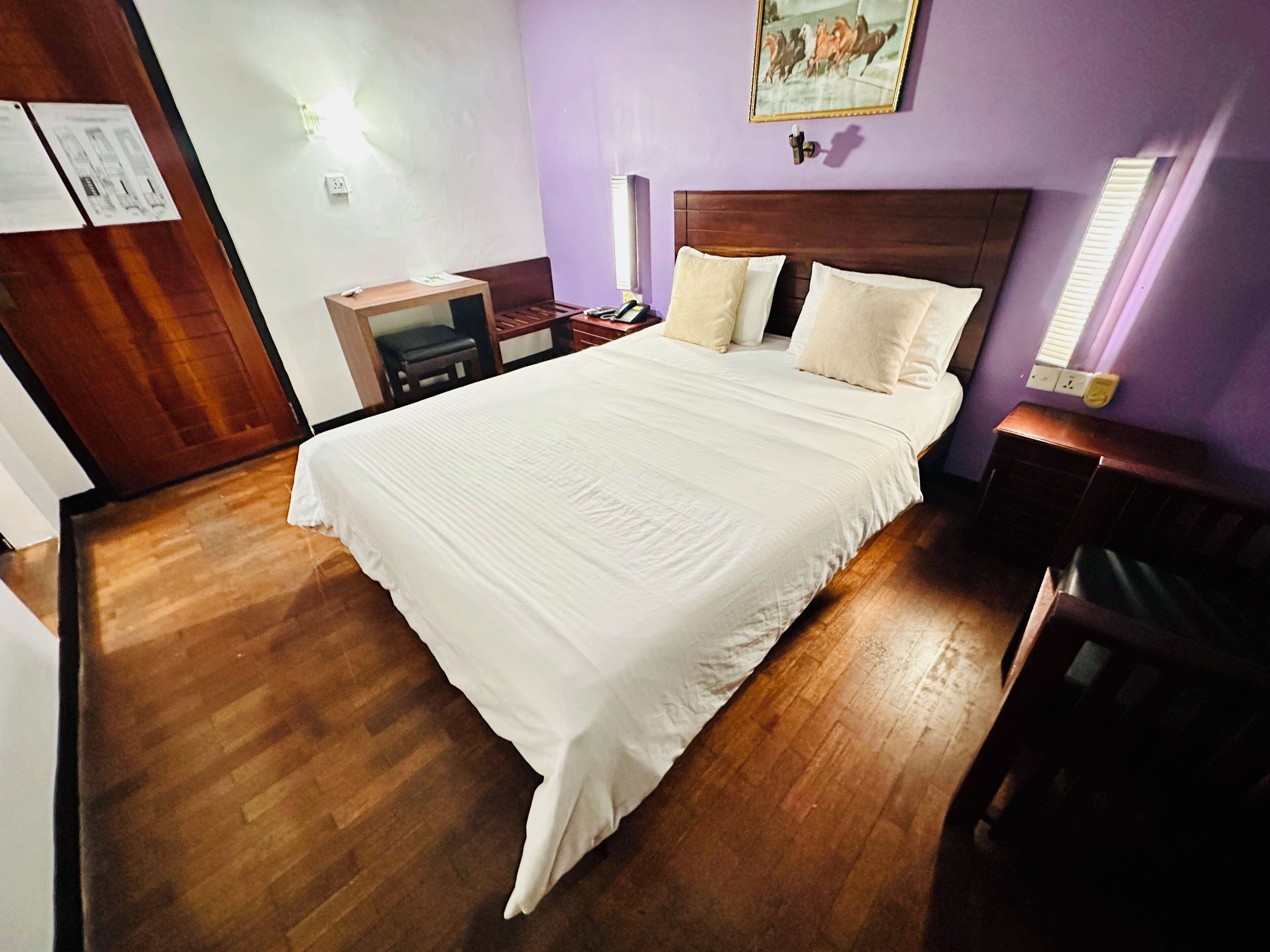 A hotel room with a large bed, wooden flooring, a desk with a chair, and a nightstand.