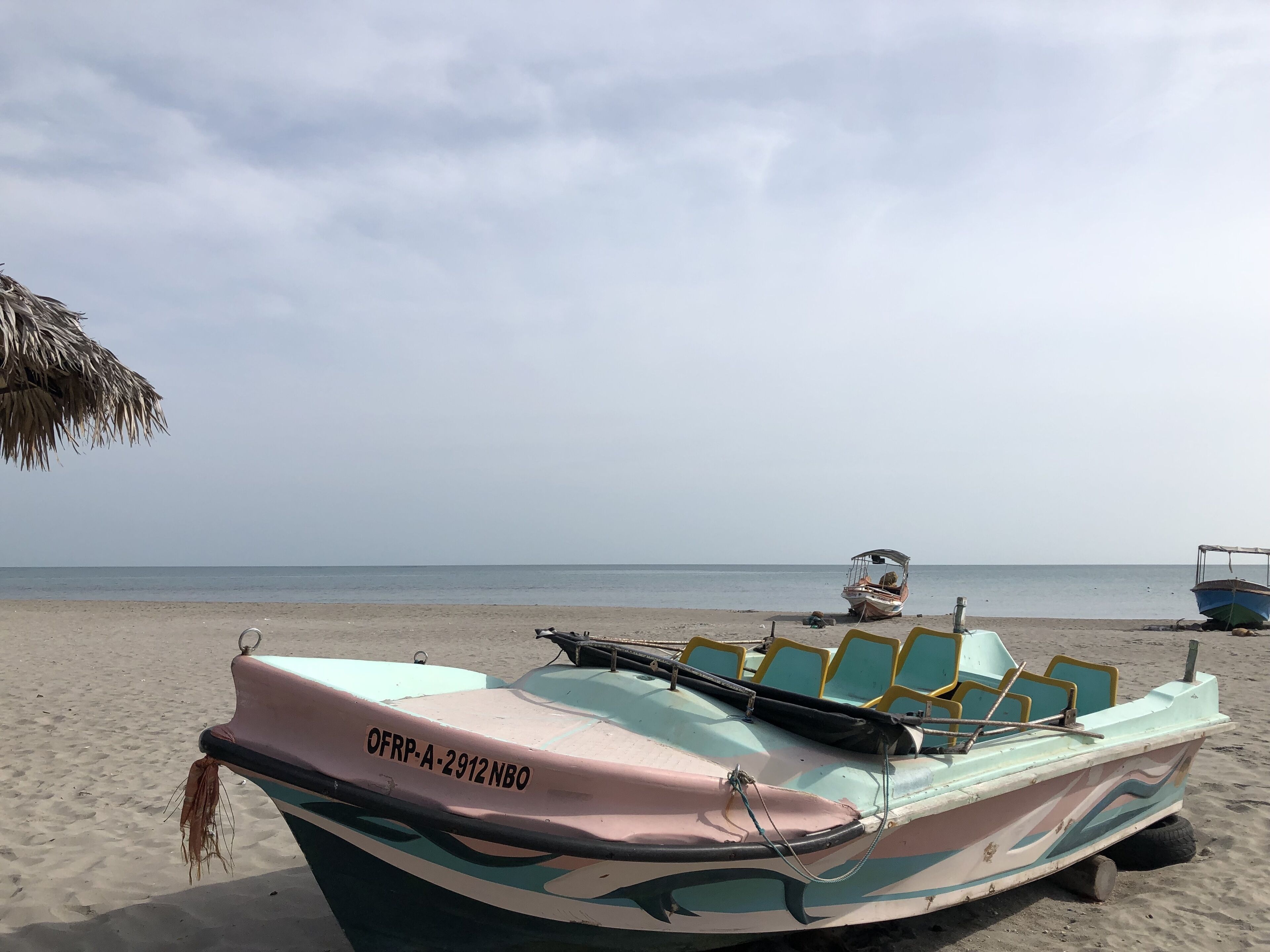 Ein Strand mit einem Ruderboot, das mit "OFRP-A-2912NBO" gekennzeichnet ist, und einem Reetdachschirm.