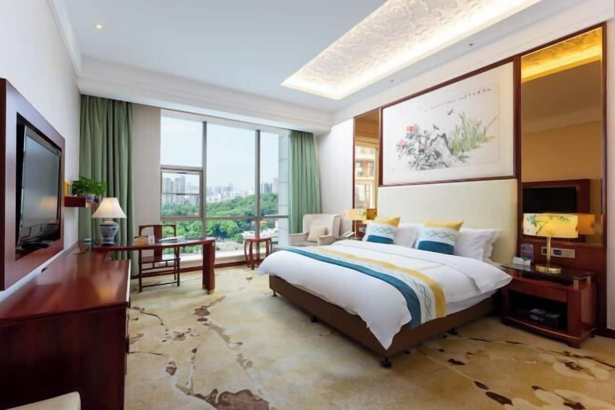 Ein Hotelzimmer mit einem großen Bett, einem Schreibtisch, einem Sessel, einem Fernseher und Blick auf die Stadt.