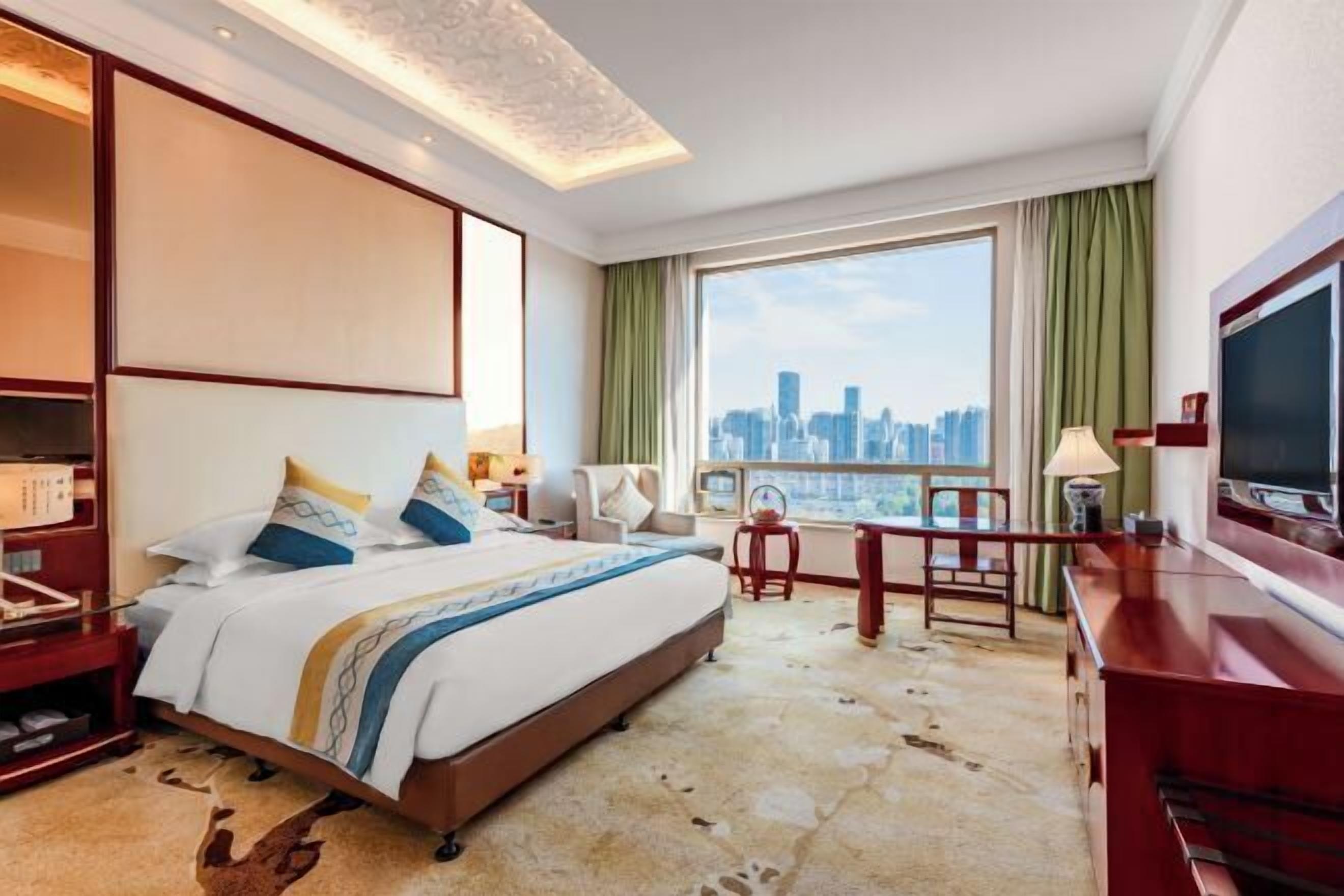 Quarto de hotel com cama, escrivaninha, cadeira, televisão e uma janela grande com vista para a cidade.