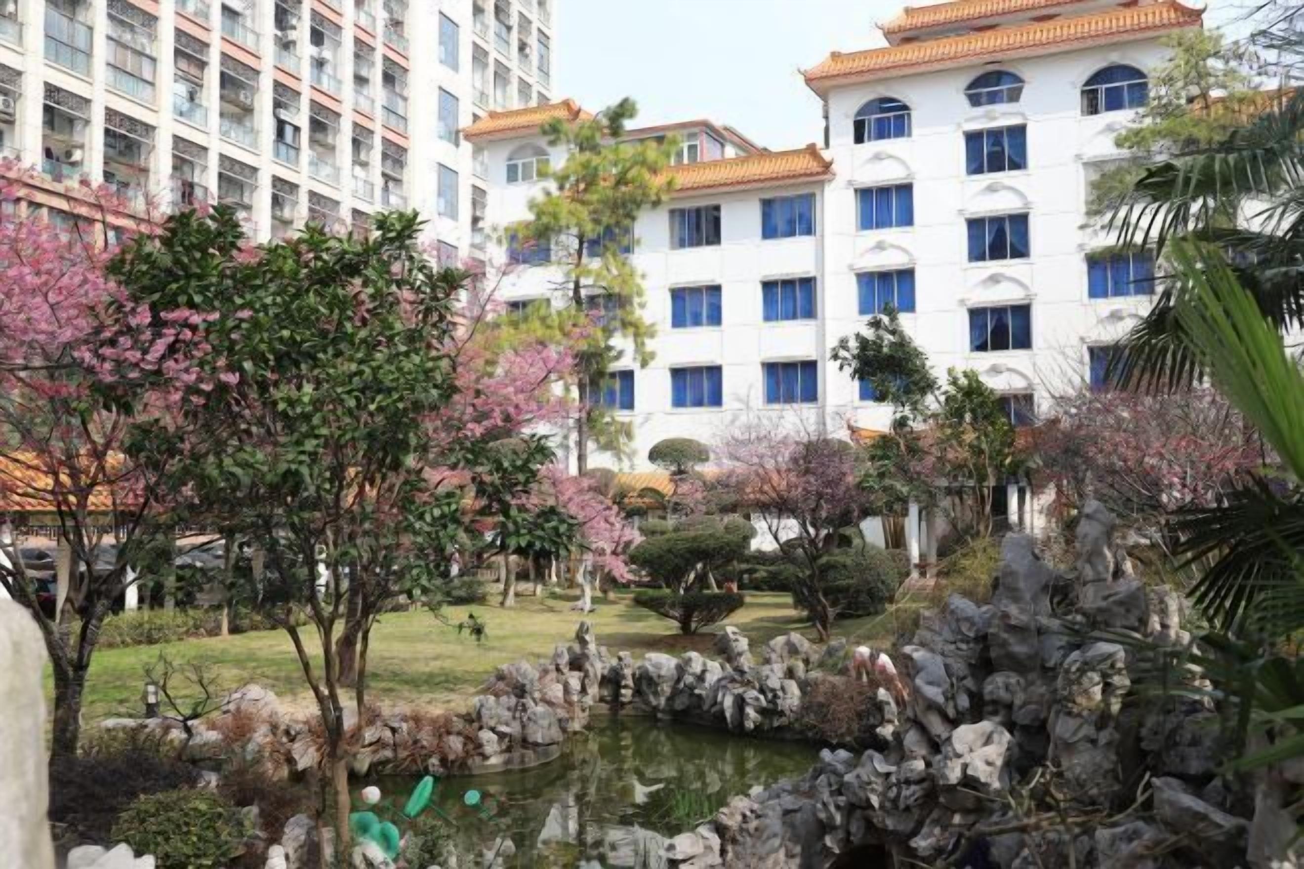 Un hôtel de style architectural traditionnel, comprenant plusieurs étages et une cour intérieure avec un bassin, entourée de divers arbres et arbustes.