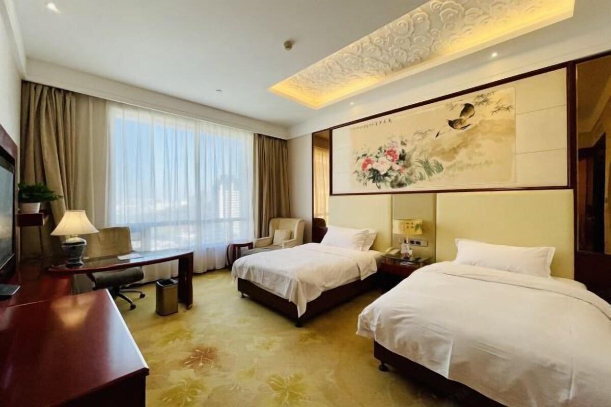 A hotel room with two beds, a desk, a chair, a TV, and a painting on the wall.