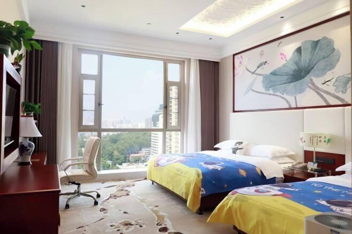 Ein Hotelzimmer mit zwei Betten, einem Schreibtisch, einem Stuhl, einem großen Fenster mit Blick auf die Stadt und einem Bild an der Wand.