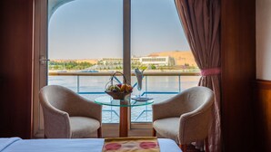 A room with a glass table, two chairs, a bed, and a view of a river and buildings. - MS Nephtis Nile Cruise (Luxor)