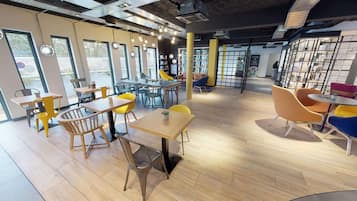 Un café moderno con mesas y sillas de madera, amplios ventanales y una zona con estanterías.