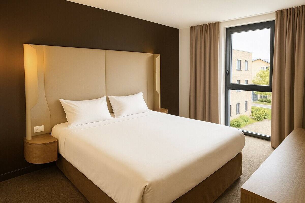 A hotel room with a large bed, a nightstand, a lamp, a window with curtains, and a view of buildings outside.