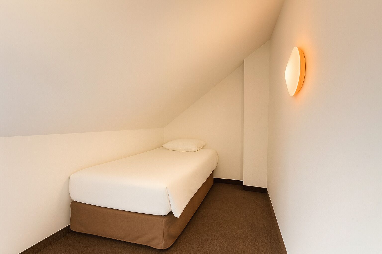 A small, single-bed room with a sloped ceiling, a wall-mounted light fixture, and a brown carpet.