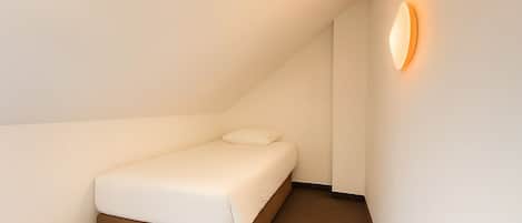 Una habitación pequeña con cama individual, techo inclinado, lámpara fijada a la pared y alfombra marrón.